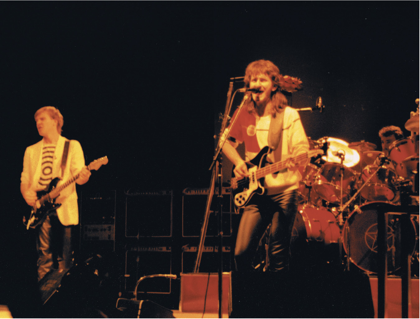 Alex, Geddy, and Neil performing on stage. To the left, Alex is playing guitar. Geddy is at the front in the centre of the picture playing guitar and singing into a microphone. Neil, who is playing the drums, is partially obscured by the drum kit.