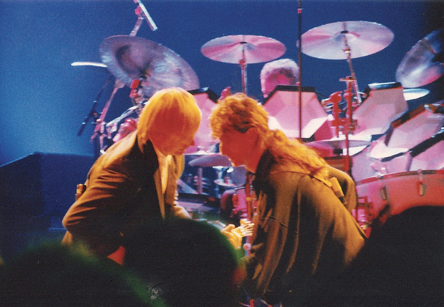 Alex and Geddy are rocking out, illuminated by an orange on-stage light.