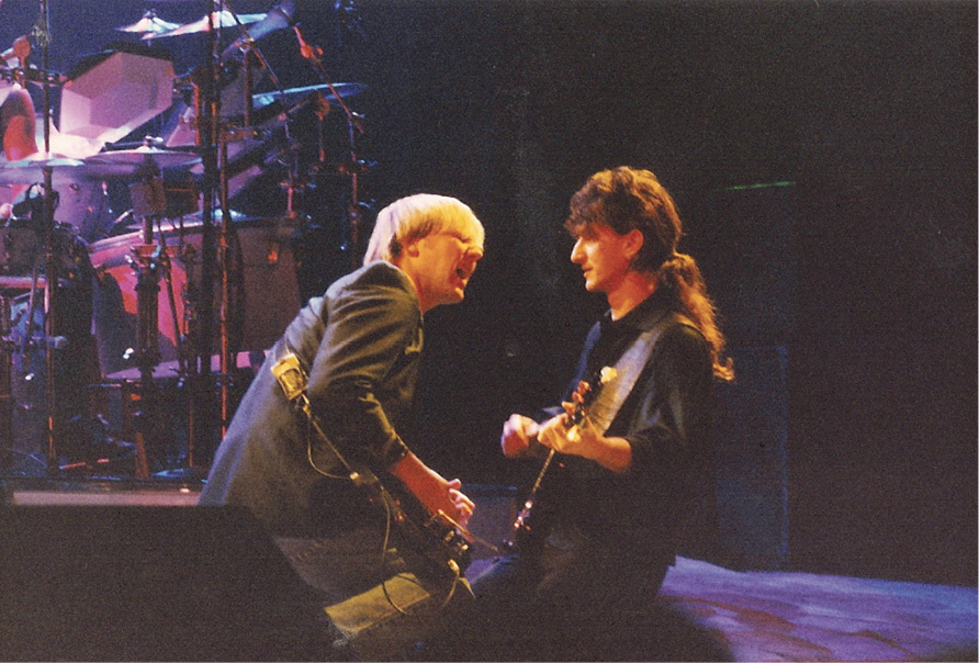 Alex and Geddy perform on stage facing each other. They're both playing the guitar within arm's length of each other.