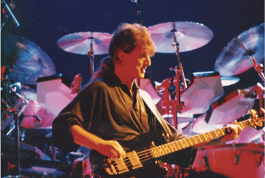 Geddy performing on a black bass guitar on stage in front of a red drum kit with cymbals.