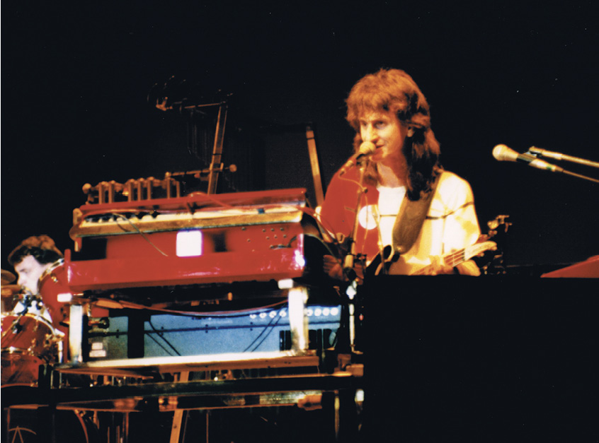 Geddy sings into a microphone as he stands behind a bank of keyboards.