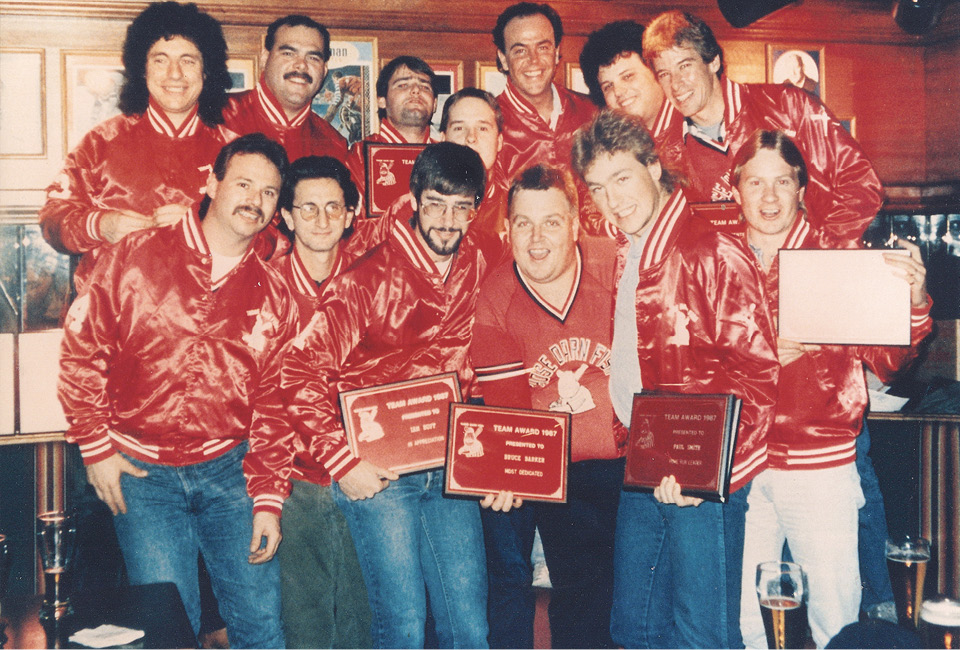 Geddy and twelve other men, all wearing matching shiny red jackets and jeans, pose for a group portrait. Four of the men at the front hold up red plaques.