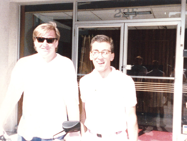 Alex, in a white shirt and straddling a bicycle, poses for a picture with Ray. Both of them are smiling.