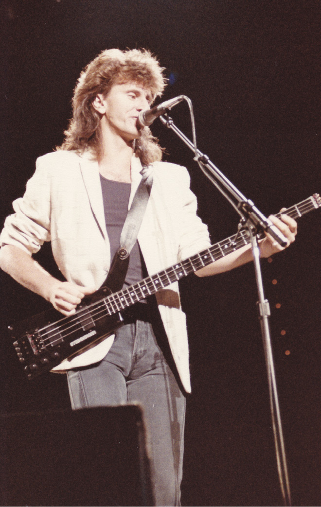 Geddy, wearing a white sports jacket, sings into the microphone as he plays a Wal bass.