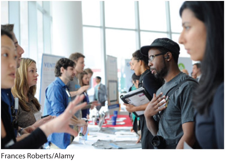 A photo shows several people seeking information from attendants standing opposite them across a table, at an event.