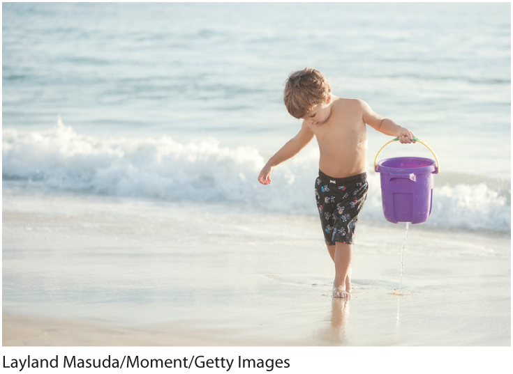 A photo shows a small boy walking alongside a beach looking down as water drips from the leaky water bucket he is holding.