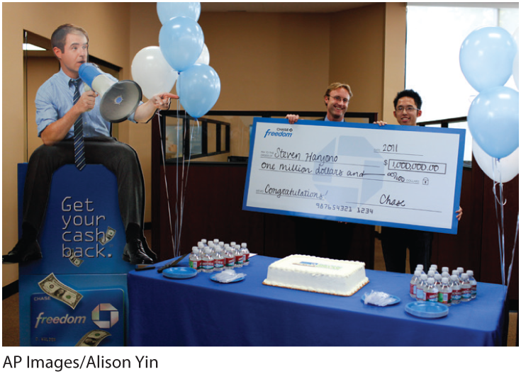 A photo shows a person receiving a lottery check of one million dollars, and a table in front of them has a cake, water bottles, and plates. Balloons and a poster depicting a man pointing and announcing through a megaphone decorate the room.