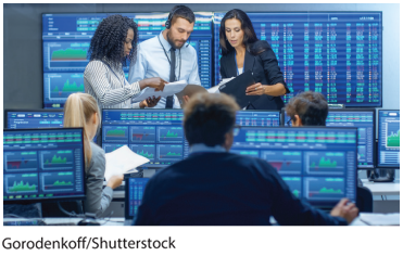 A photo shows people working in a stock market office, looking at their computers displaying graphs. Three of them are analyzing the sheets in their hands.