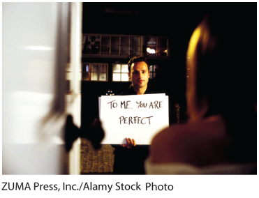 A photo of a man showing a sign that reads, ‘To me you are perfect’ to a woman standing near a door.