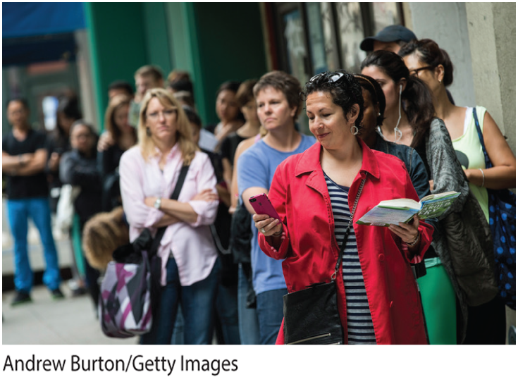 A photo shows people waiting in a line. The lady in the front has an open book in one hand and a mobile phone in the other hand.