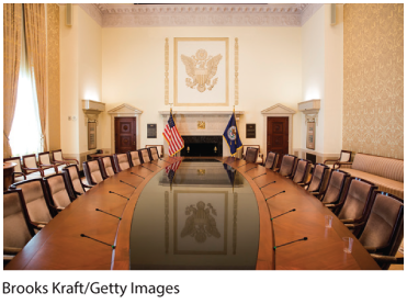 A photo shows the Federal Reserve Systems board room. Chairs line either side of a long table set fitted with microphones, the Federal Reserve Systems Flag, and the United States national flag at the head.