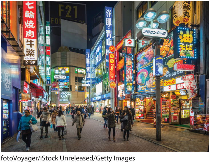 A photo shows people walking by a cobbled street with bright neon signs in Japanese. 