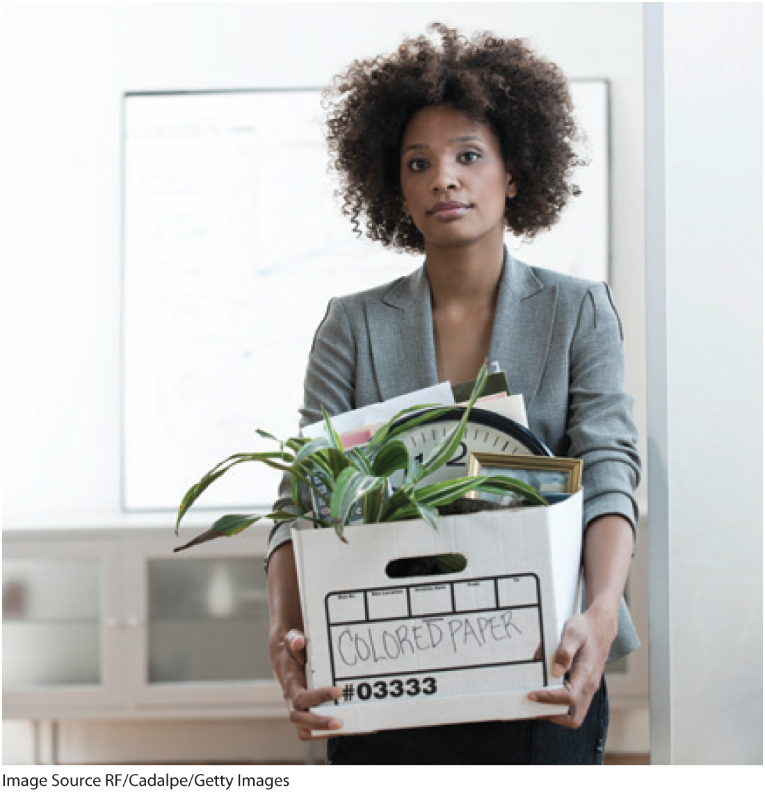 A photo shows a woman with a box containing a potted plant, photo frame, and papers.