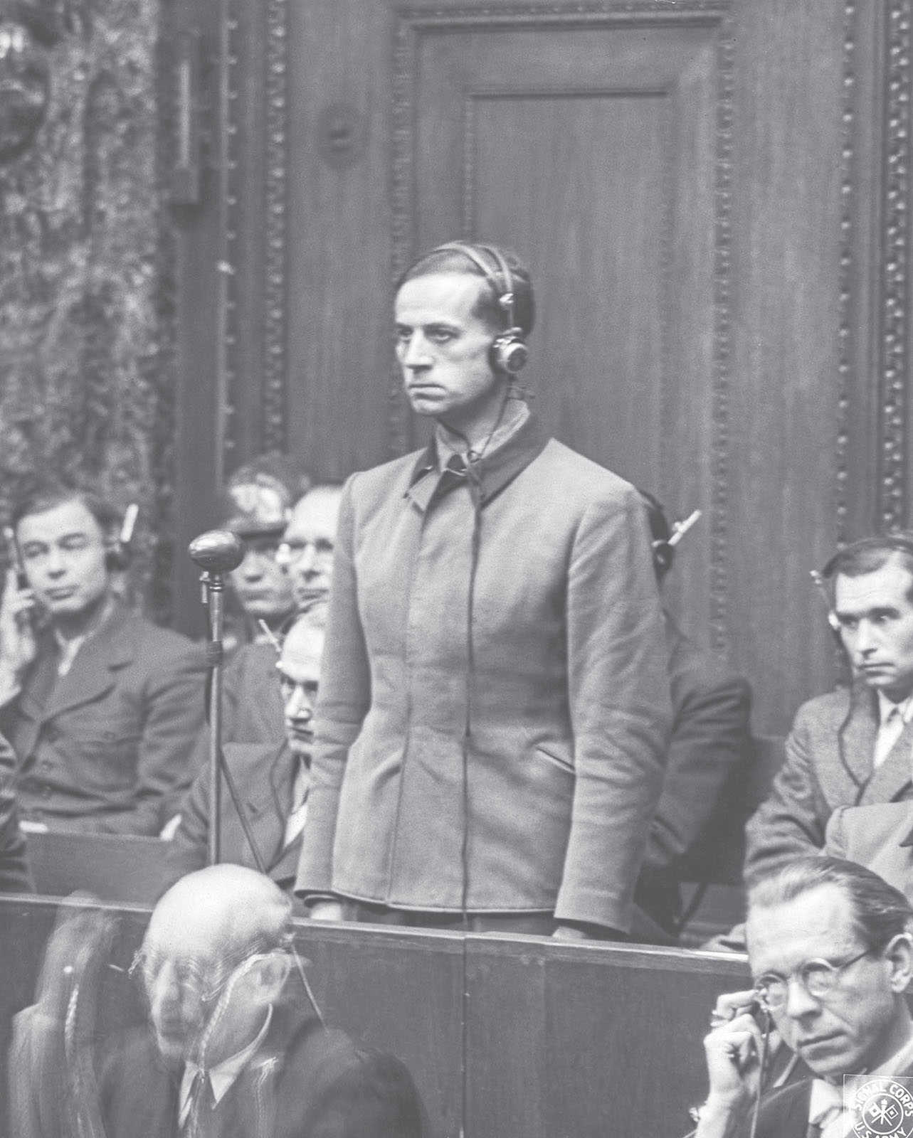 Karl Brandt wears headphones and stands in a courtroom during his trial. Other men in the courtroom sit behind him with headphones on.